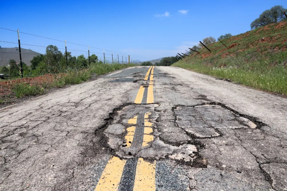 Close-up of cracked and damaged two-lane roadway with double yellow stripes on a bright sunny day.